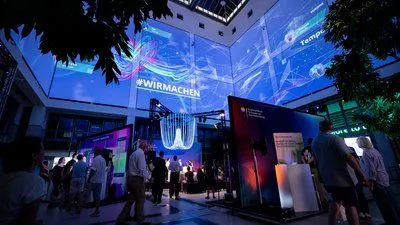 Indoor event space with several exhibition booths and visitors, featuring large digital projections overhead with the text “#WIRMACHEN”
