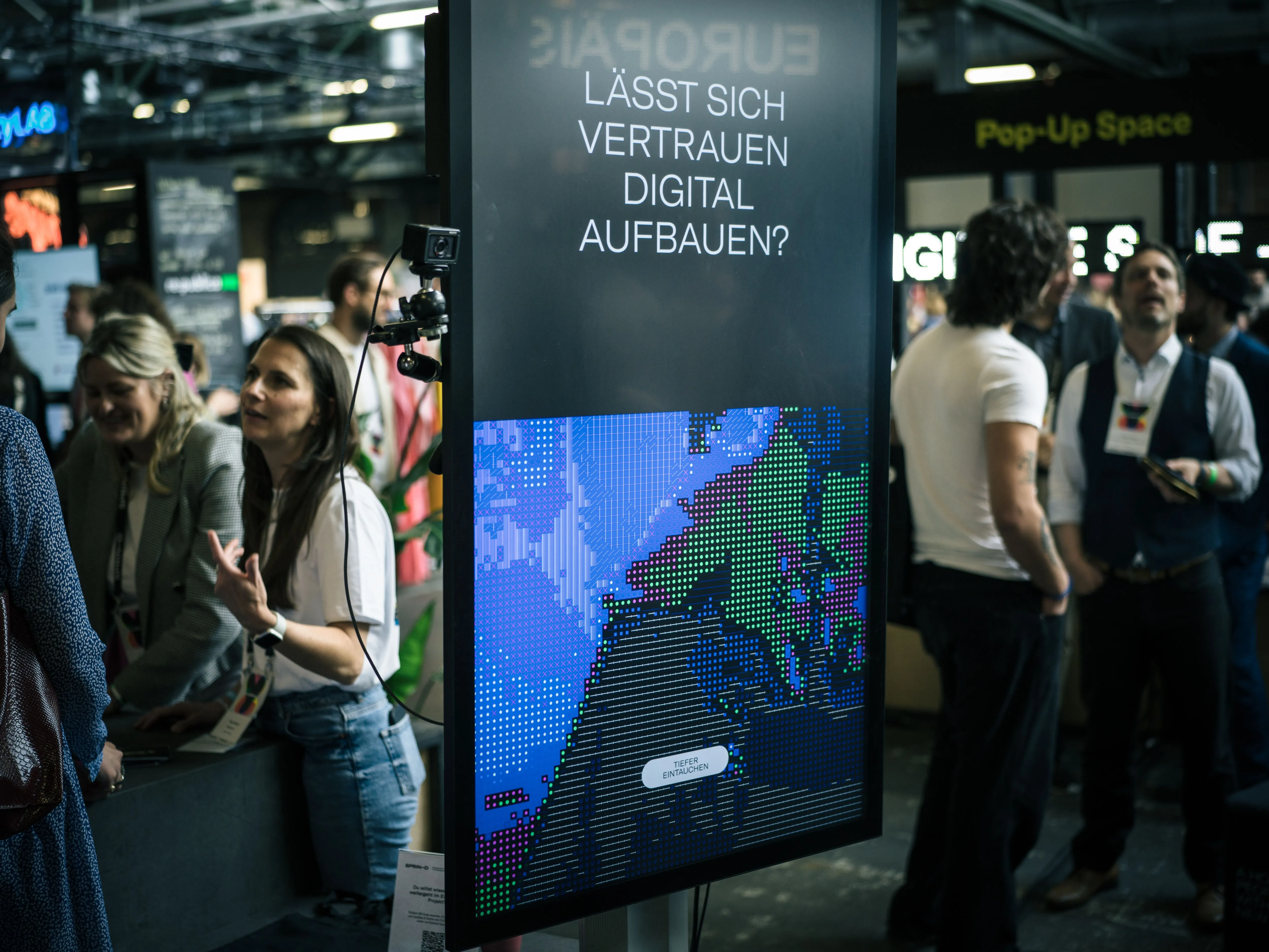 Veranstaltungsraum mit mehreren Personen im Gespräch. Im Vordergrund ein großes Display mit der Frage „Lässt sich Vertrauen digital aufbauen?“ und einer grafischen Darstellung