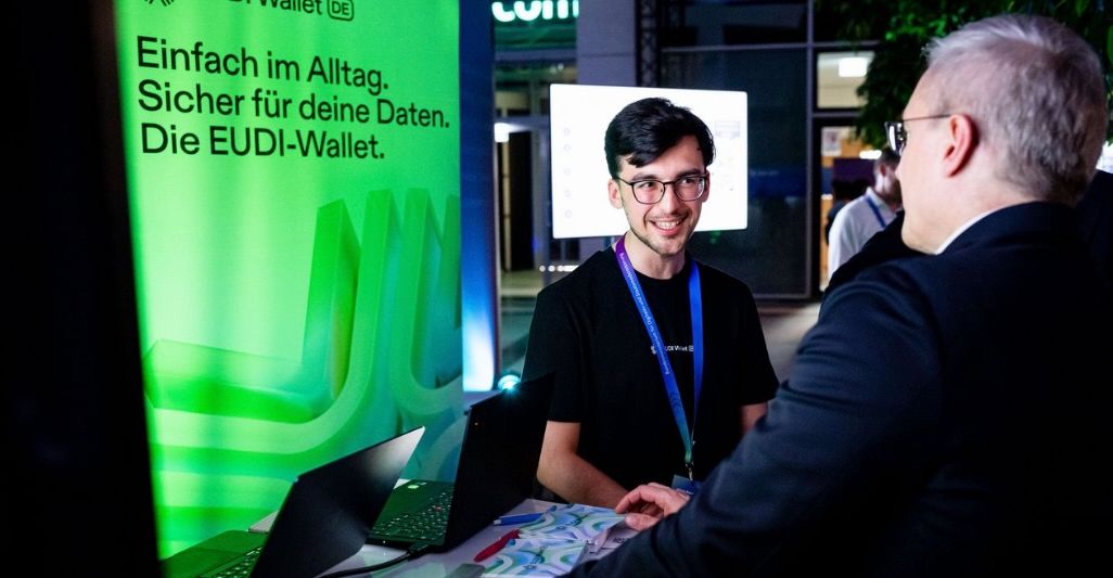 Gespräch an einem Messestand zur EUDI‑Wallet vor einer grünen Informationswand mit dem Slogan „Einfach im Alltag. Sicher für deine Daten. Die EUDI‑Wallet.“