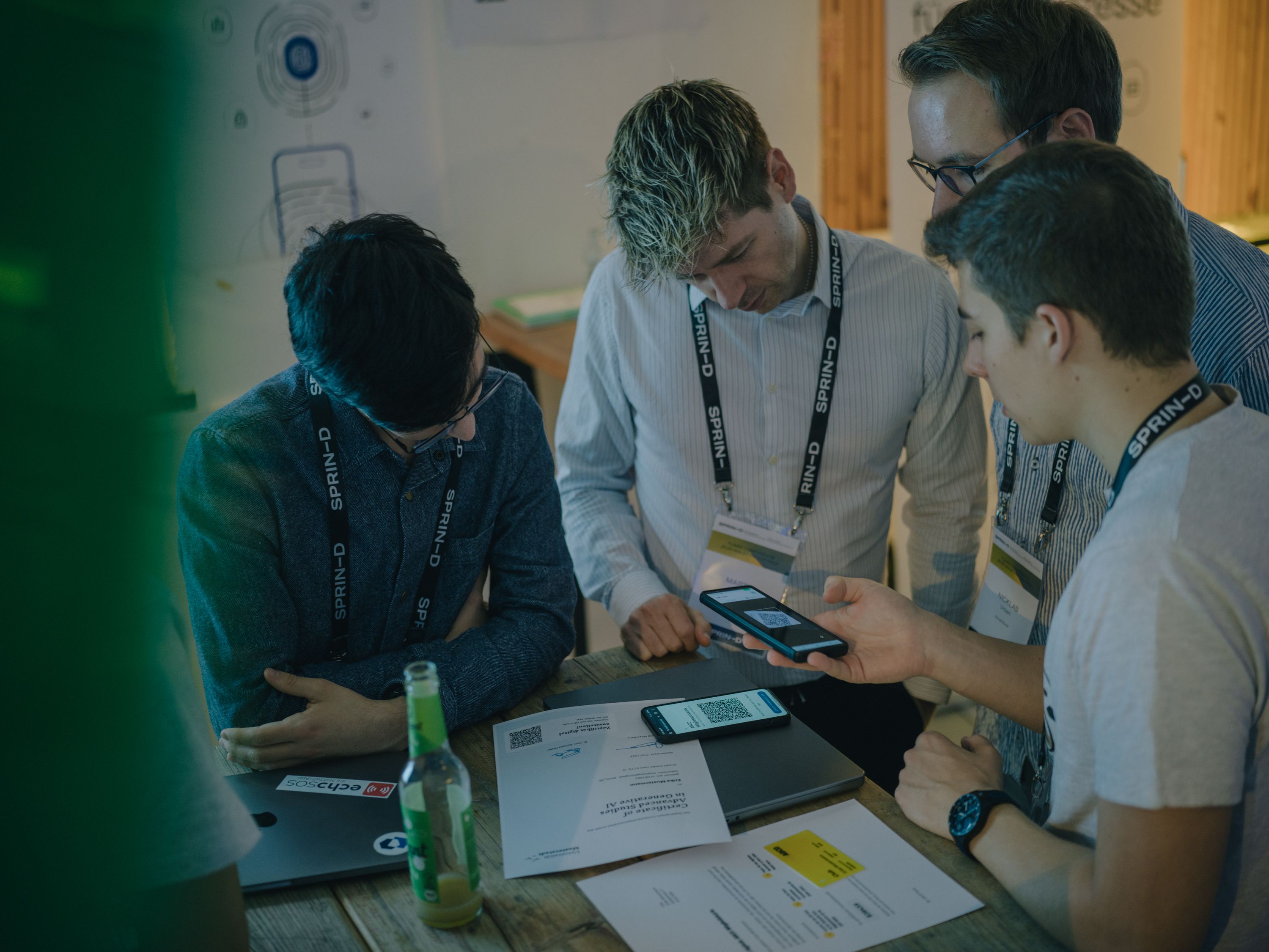 Several participants stand around a table looking at a smartphone displaying a QR code, with documents and laptops placed on the table