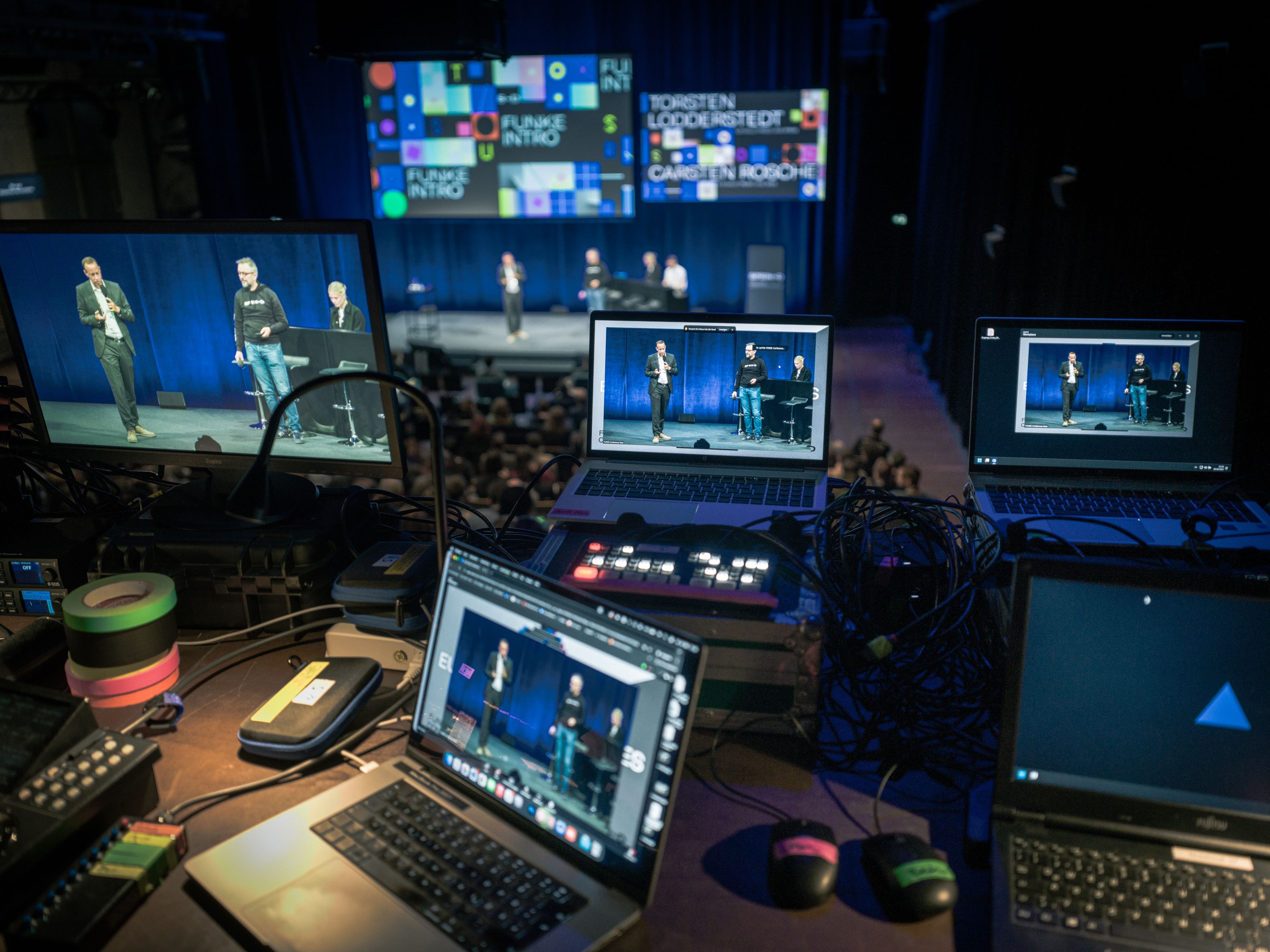 Picture of a dark video production control room equipped with numerous laptops and monitors. All screens are displaying a live event from the perspective of the control desk.