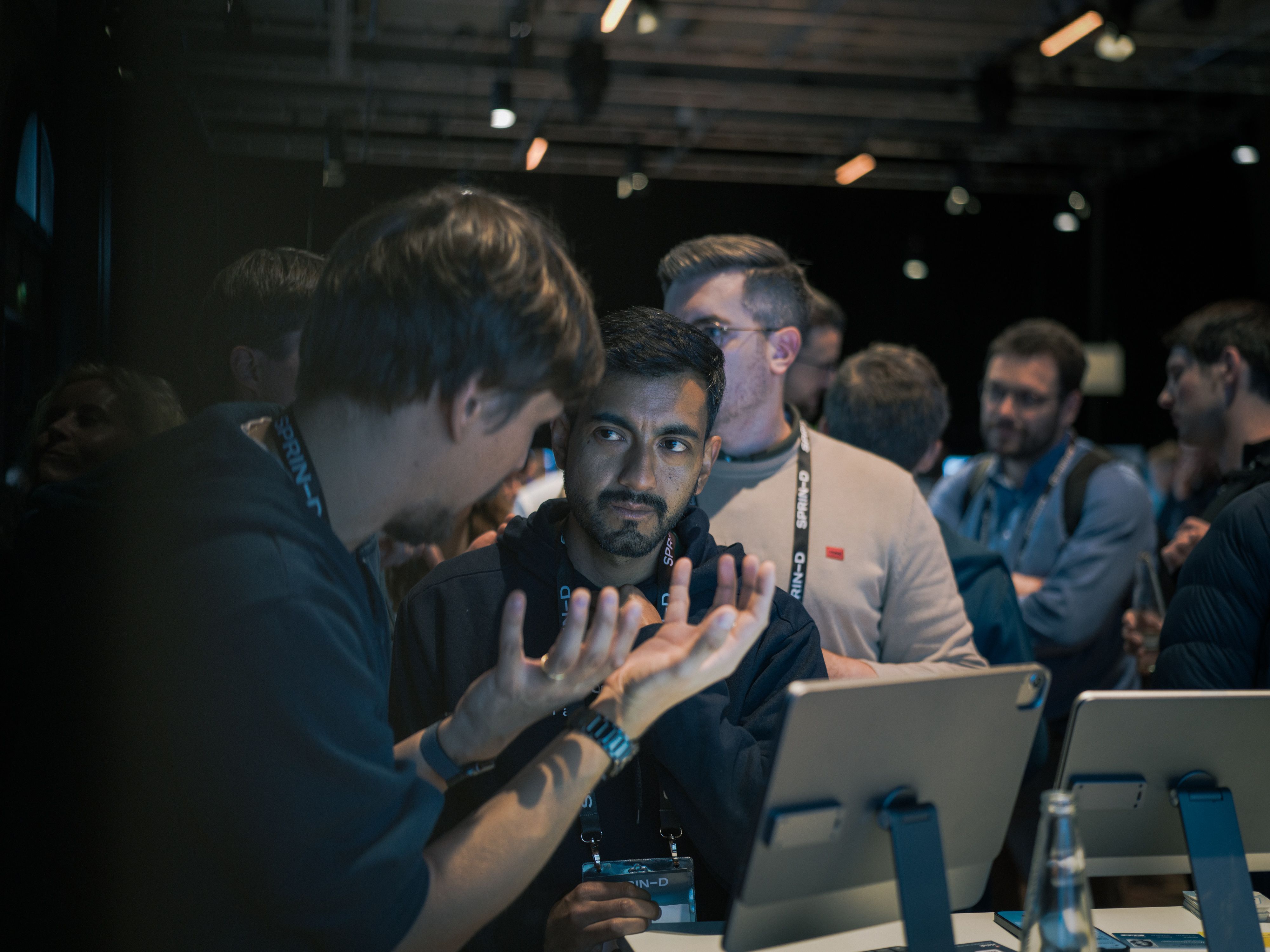 Two men talk intently at a table with tablets, surrounded by people at an event.