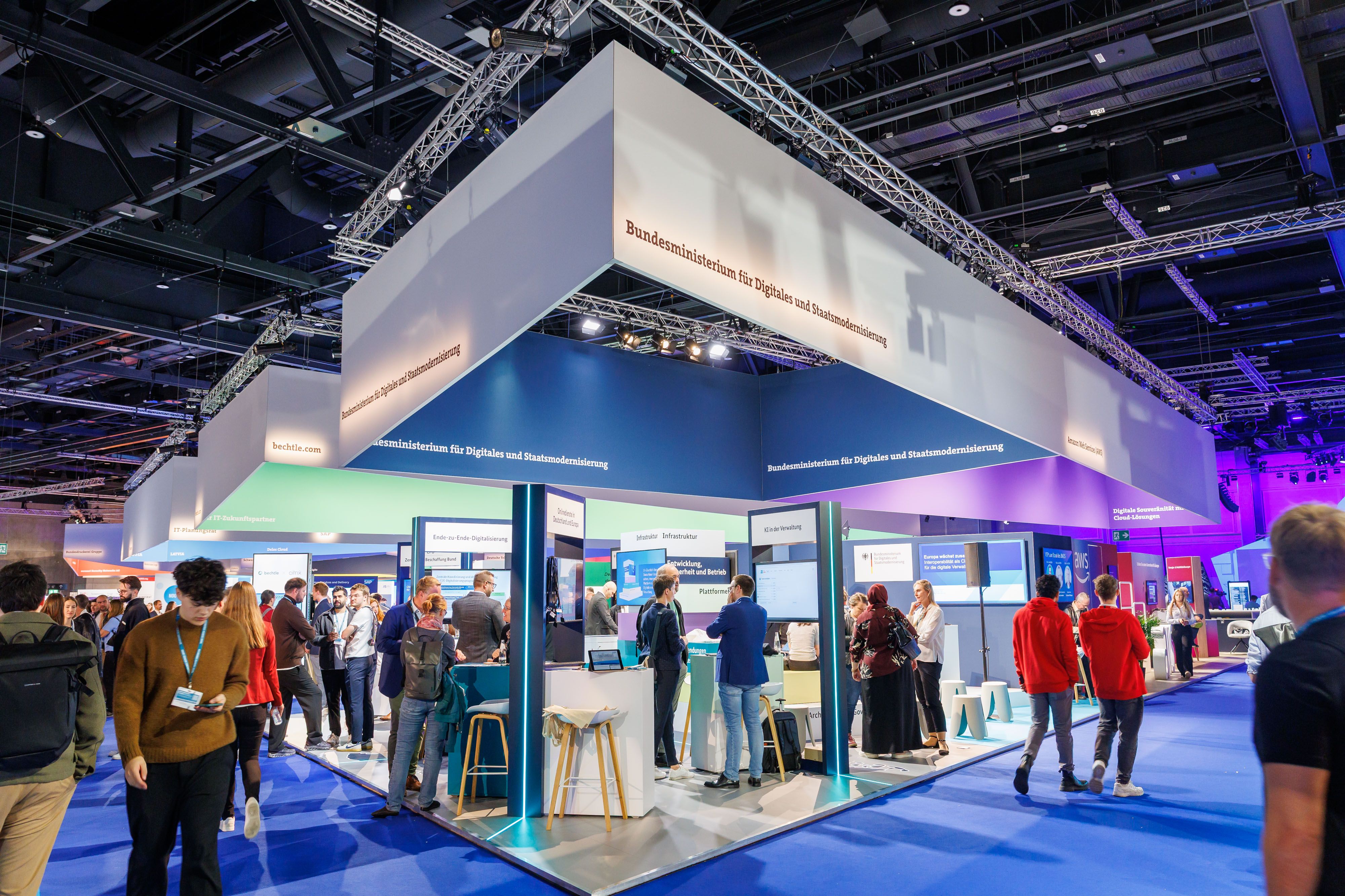Stand des Bundesministeriums für Digitales und Staatsmodernisierung auf einer Messe, mit mehreren Personen im Gespräch und digitalen Präsentationen.