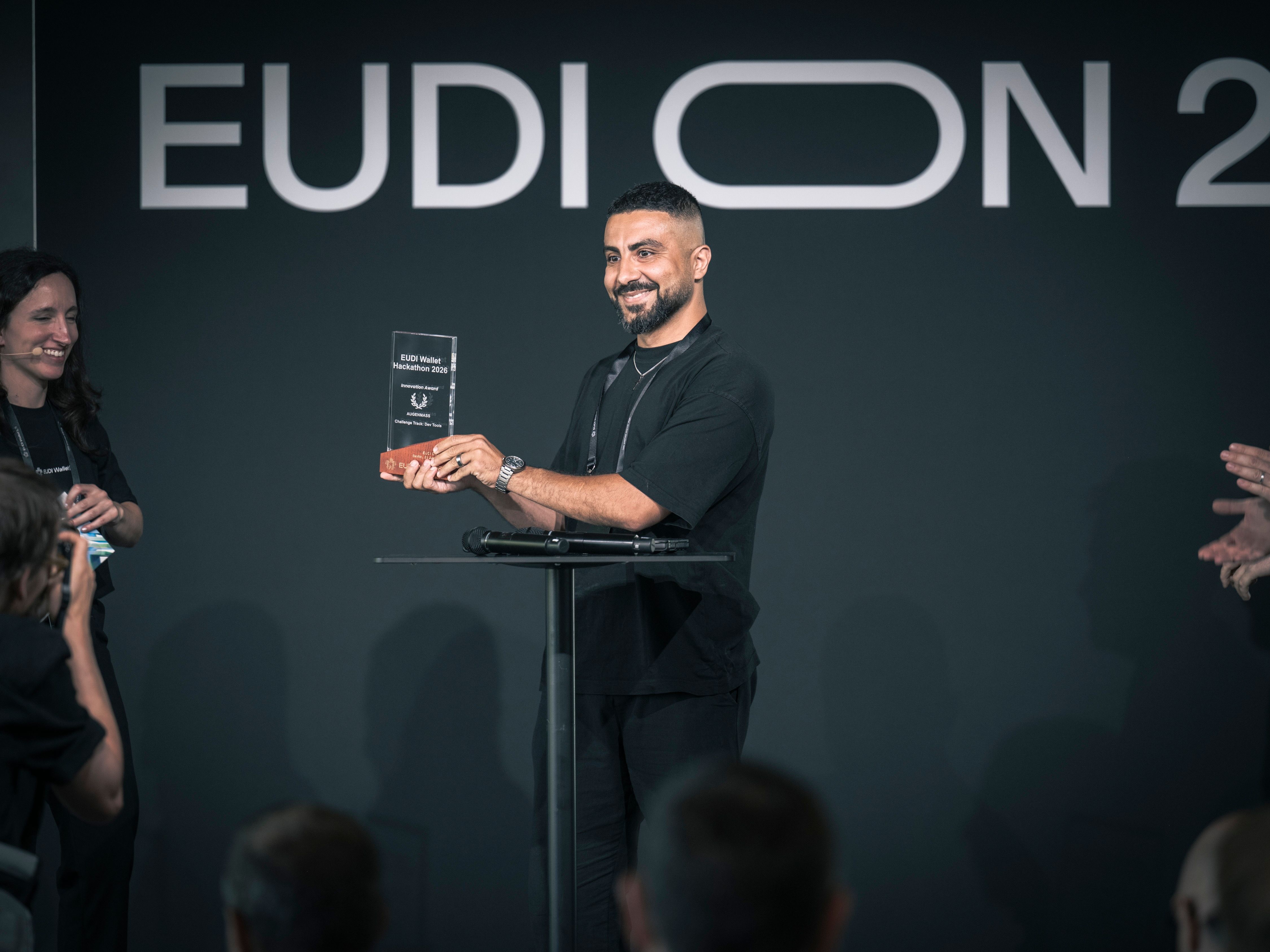  A smiling man holds up a glass "EUDI Wallet Hackathon 2026" award on stage at EUDI ON 2026 while people beside him applaud.