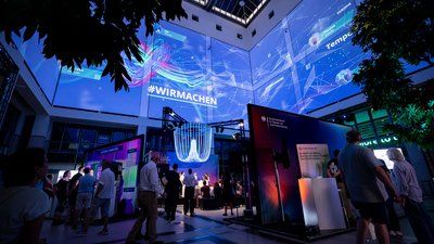 Innenaufnahme eines Veranstaltungsbereichs mit mehreren Messeständen und Besucherinnen und Besuchern, über denen große digitale Projektionen mit der Aufschrift „#WIRMACHEN“ zu sehen sind
